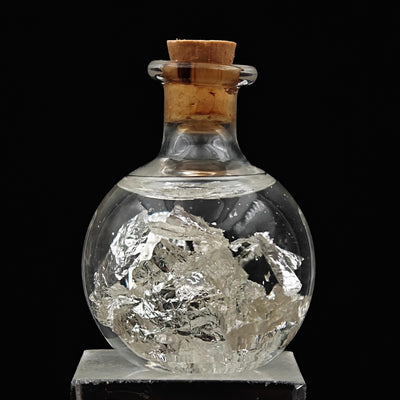 Glass vial with delicate natural silver leaf flakes, front view on stand